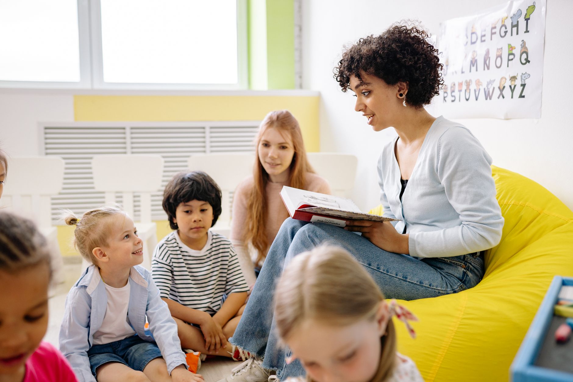 Maestra joven leyendo un cuento a un grupo de niños sentados en el suelo de un aula de infantil